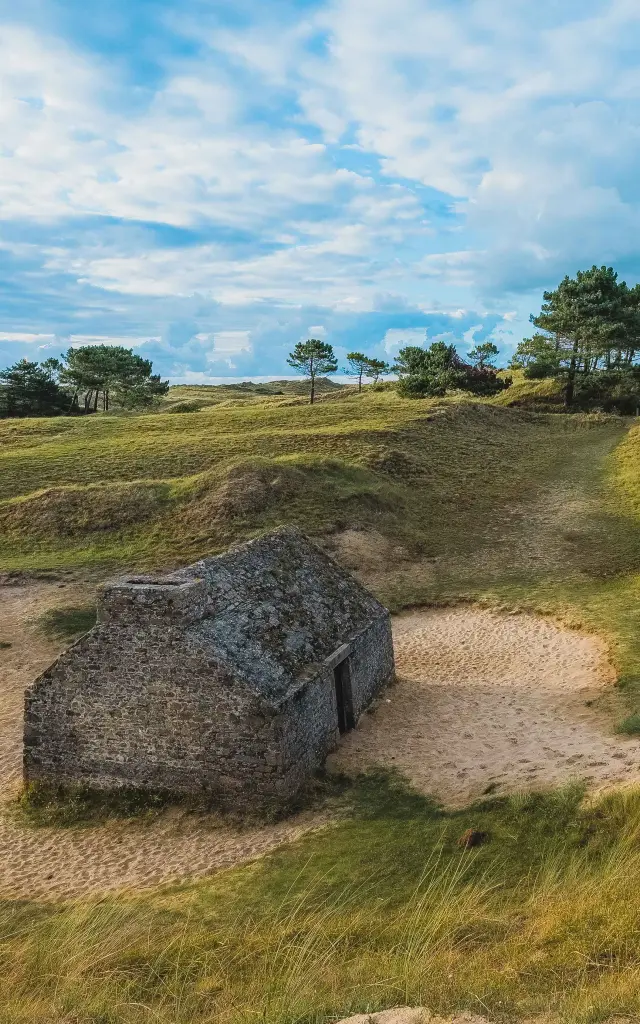 Cabane de douanier en pierre entouré de dunes herbeuses et de quelques arbres
