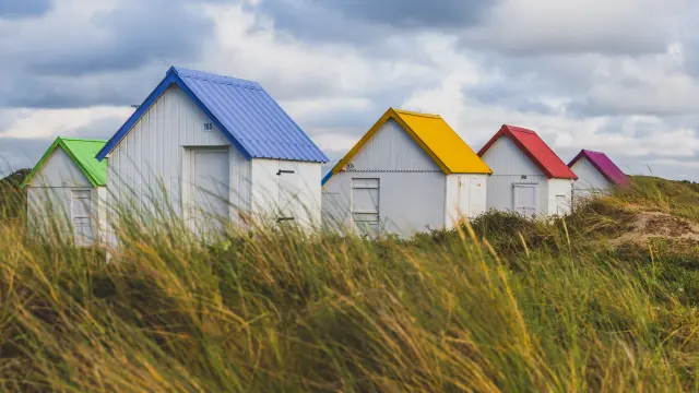 Trois maisons de pêcheur alignées avec toits bleus et façades blanches, entourées de hautes herbes