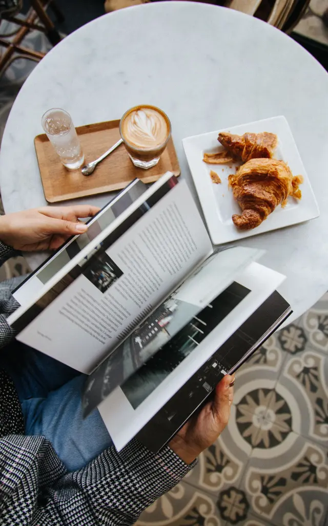 Personne lisant un magazine à une table avec un café et des croissants