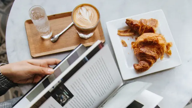 Personne lisant un magazine à une table avec un café et des croissants