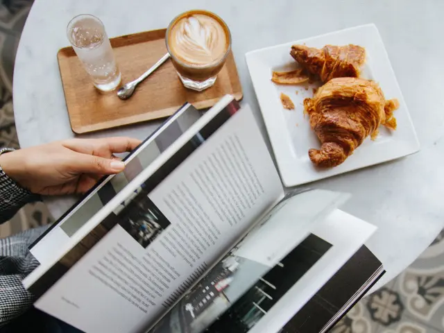 Personne lisant un magazine à une table avec un café et des croissants