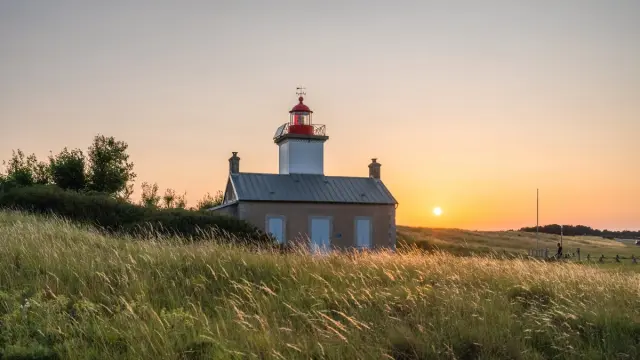 Phare en pierre avec une lanterne rouge et blanche, entouré de hautes herbes, au coucher du soleil