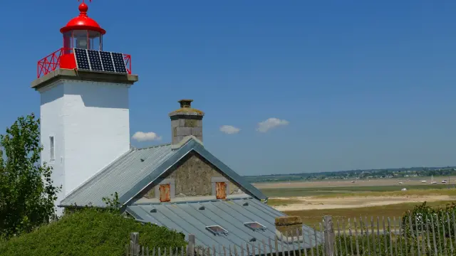 White lighthouse with a red lantern and a solar panel, adjacent to a gray house with a metal roof