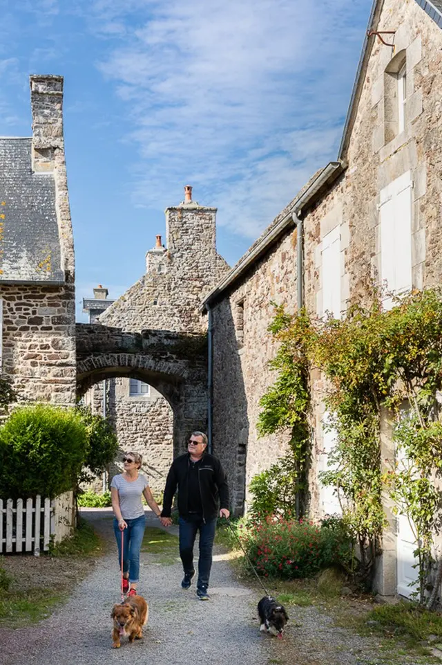 Deux personnes marchant avec deux chiens dans une allée entre des bâtiments en pierre à Regnéville sur Mer