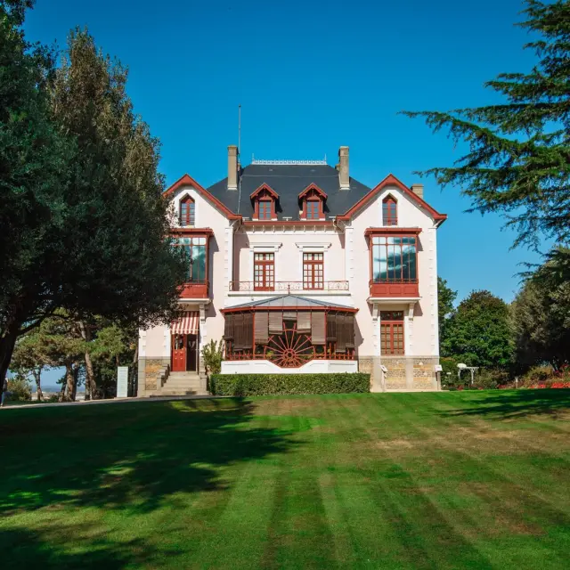 Two-story Victorian manor with red brick facade and white trims, surrounded by green lawn and trees