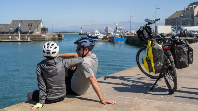 Motorcyclist in protective gear sitting on a bench facing a harbor with sailboats