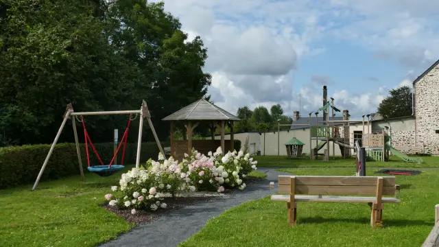 Aire de jeux en bois avec toboggans rouges et balançoires, entourée de fleurs blanches et de gazon
