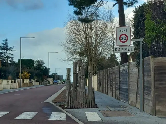 Panneau de signalisation indiquant une zone limitée à 30 km/h avec rappel, situé près d’une route bordée d’arbres et de bâtiments