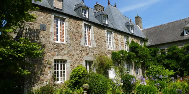 Manoir en pierre avec des fenêtres à petits carreaux, entouré de végétation, situé près d’un plan d’eau