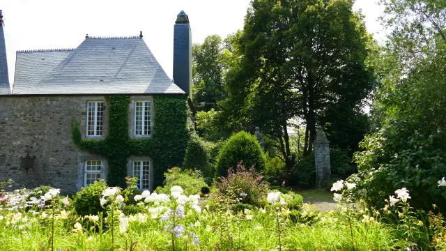 Maison en pierre avec volets verts et cheminée, entourée de fleurs et d’arbres