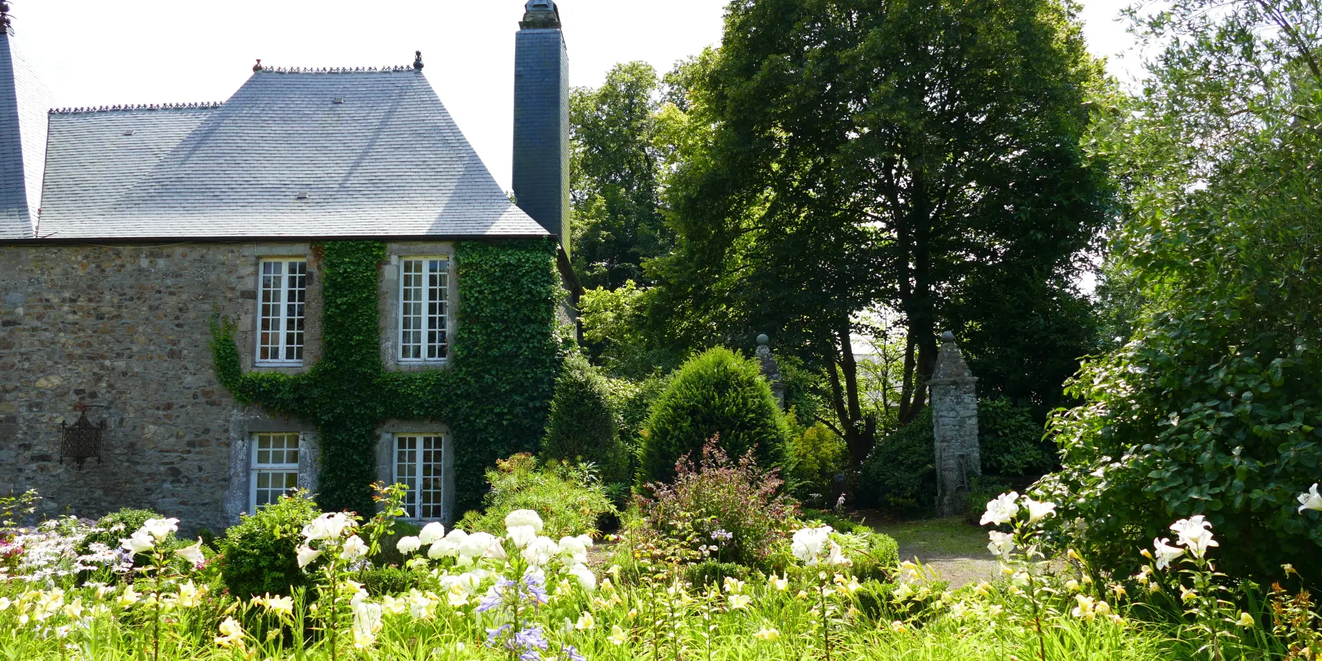 Maison en pierre avec volets verts et cheminée, entourée de fleurs et d’arbres