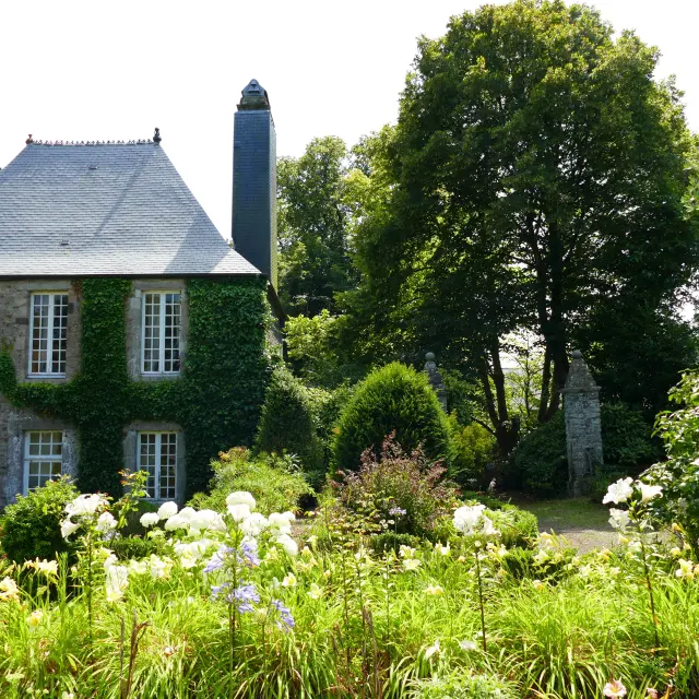 Maison en pierre avec volets verts et cheminée, entourée de fleurs et d’arbres