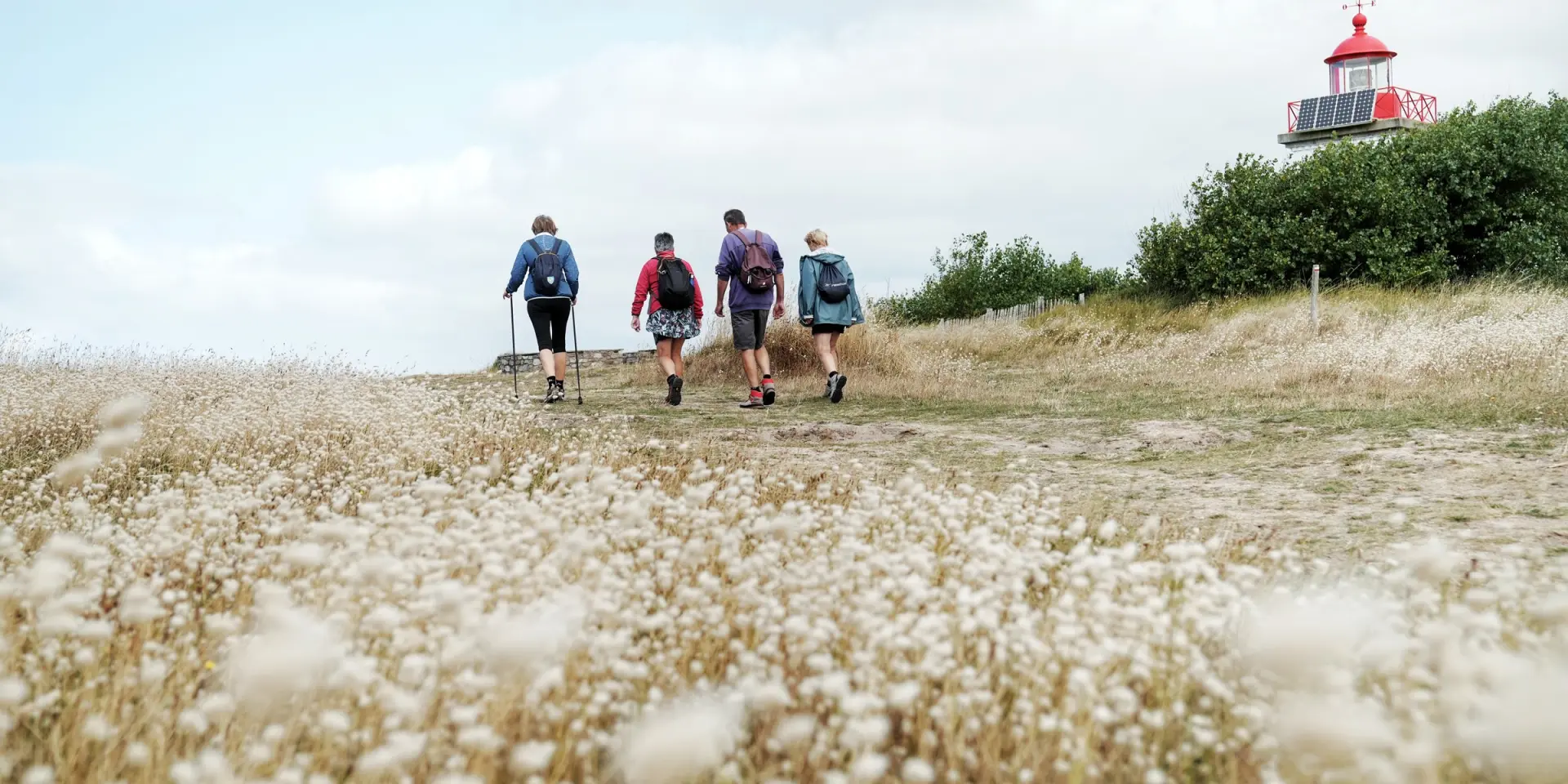 Quatre personnes marchant vers un petit phare rouge et blanc dans un champ d'herbes hautes