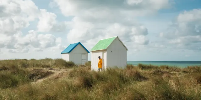 Deux cabines de plage blanches avec des toits colorés, l'une verte et l'autre bleue, sur une dune herbeuse près de la mer.- Gouville sur Mer