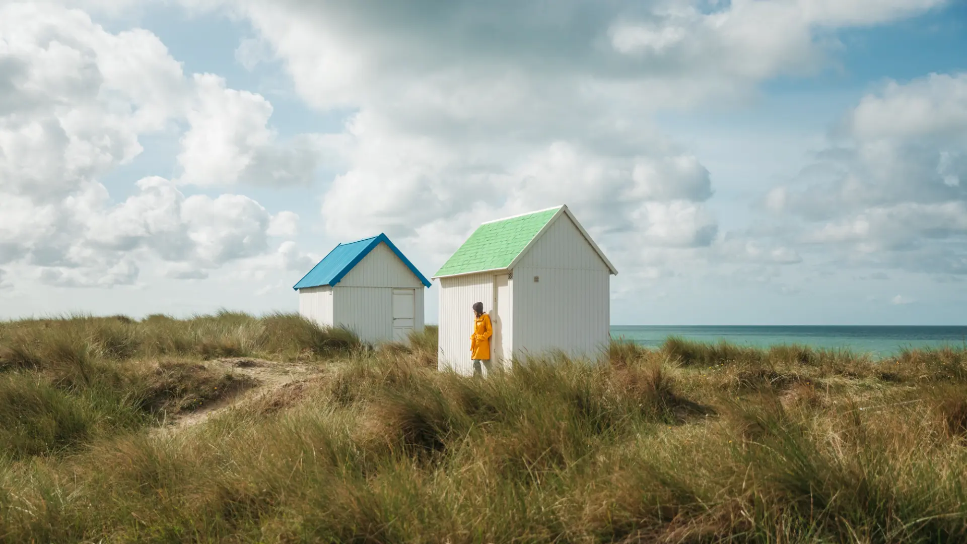 Zwei weiße Strandhütten mit bunten Dächern, eine grüne und eine blaue, auf einer grasbewachsenen Düne in der Nähe des Meeres.