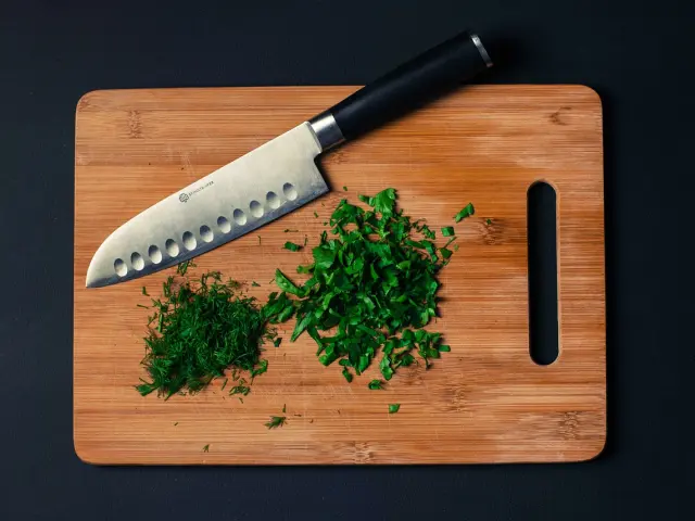 Un couteau de chef posé sur une planche à découper en bois avec des herbes vertes hachées