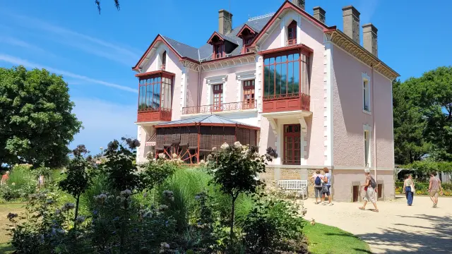 Multi-story brick house with red and white facade and shuttered windows, surrounded by greenery and pedestrians