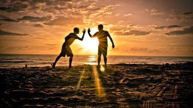 Deux personnes marchant sur une plage au coucher du soleil, avec des voiliers amarrés en arrière-plan