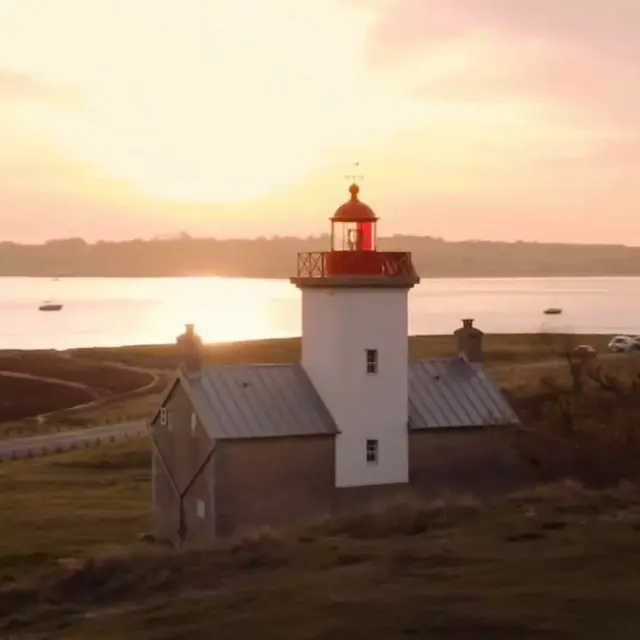 Phare blanc avec une bande rouge au sommet, entouré de bâtiments bas et de voiliers amarrés au coucher du soleil