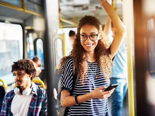 Young gorgeous cheerful woman is standing on the bus using the phone and smiling.