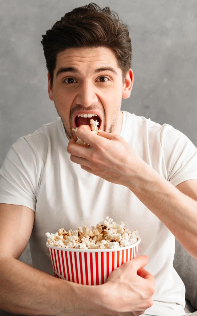 Single man 20s sitting on sofa in gray apartment and eating pop corn, while watching tv in morning