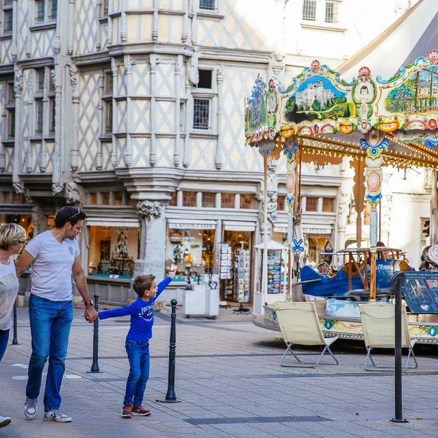 Un couple et un enfant passant sur la place, l'enfant montre le manège