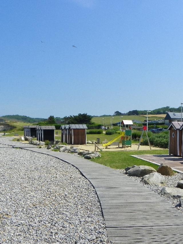 Cabines de plage en bois marron, aire de jeux et planches pour marcher sur les galets. Falaises et mer à l'arrière-plan