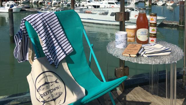 Produits de la boutique de l'Office de Tourisme (marinière, tote bag Deeptown, cidre, miel, caramels de pommes, mug, carnet, stylo) présentés sur un transat et une table avec le port de plaisance en arrière-plan.