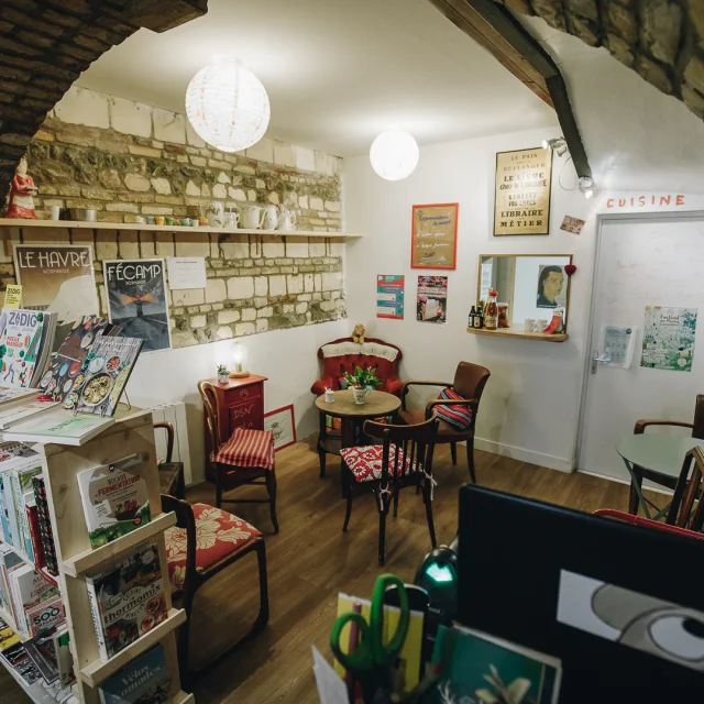 Coin goûter dans une librairie. Pièce voûtée, fauteuils dépareillés, affiches aux murs, étagère remplie de livres.