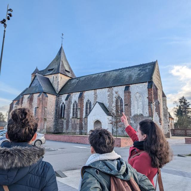 Une guide conférencière mont du doigt l'église de Saint-Nicolas d'Aliermont à deux jeunes femmes participant à la visite guidée