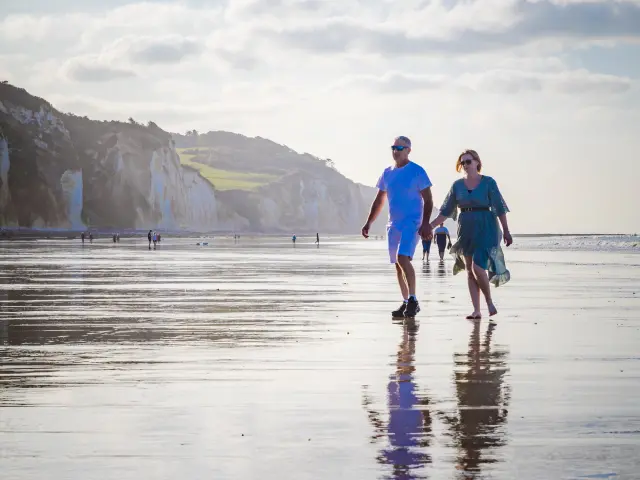 Un couple homme et femme se balade main dans la main sur la plage de Pourville à marée basse, falaises en arrière plan