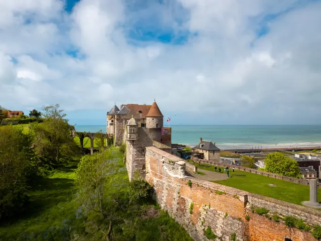 Le château de Dieppe vu depuis les airs, entouré de verdure, dominant la ville et la mer sous un ciel légèrement nuageux.