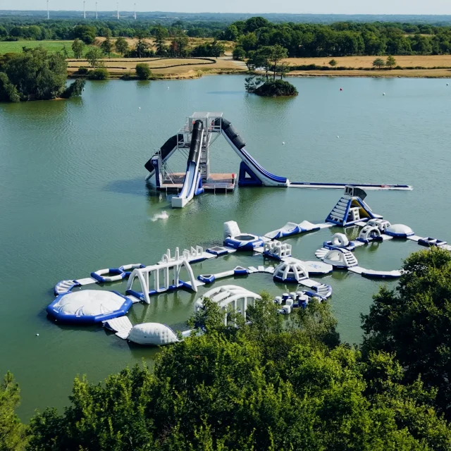 Vue aérienne de l'Espace Quilly. Vue sur le plan d'eau avec les structures gonflables flottantes bleues et blanches et sur la tour géante avec ses toboggans. En arrière plan, un paysage de campagne avec des éoliennes