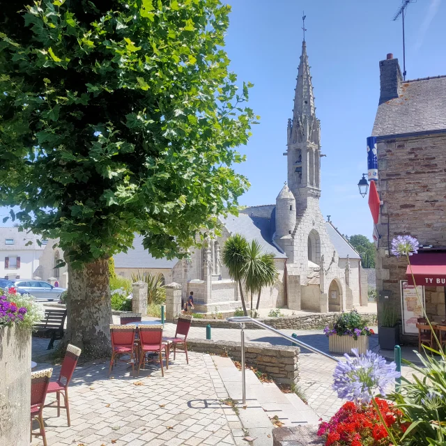 Zicht op de buitenkant van de Notre Dame Izel kerk met het terras van de Ty Butun bar en zijn platanen
