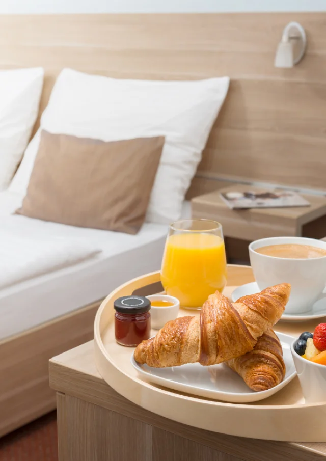 Chambre d'hôtel avec un lit double et un petit déjeuner servi avec un jus de fruits, des croissants, de la confiture, un café et un bol de fruits frais sur une petite table