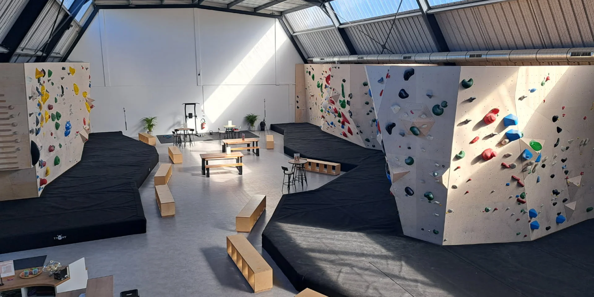 Salle d'escalade vide avec les différents murs de grimpes et leurs différents niveaux, indiqués par les couleurs noir, bleu, vert, jaune et rouge. Des matelas de réception sont disposés au bas des murs.