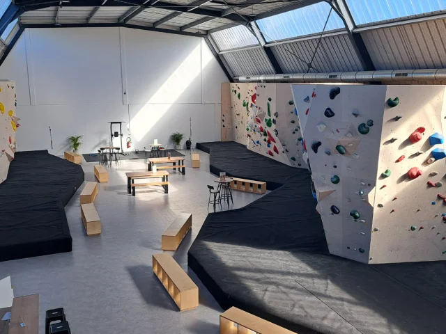 Salle d'escalade vide avec les différents murs de grimpes et leurs différents niveaux, indiqués par les couleurs noir, bleu, vert, jaune et rouge. Des matelas de réception sont disposés au bas des murs.