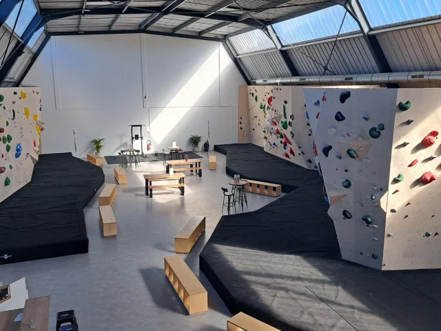 Salle d'escalade vide avec les différents murs de grimpes et leurs différents niveaux, indiqués par les couleurs noir, bleu, vert, jaune et rouge. Des matelas de réception sont disposés au bas des murs.