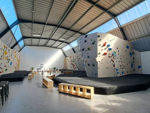 Salle d'escalade vide avec les différents murs de grimpes et leurs différents niveaux, indiqués par les couleurs noir, bleu, vert, jaune et rouge. Des matelas de réception sont disposés au bas des murs.