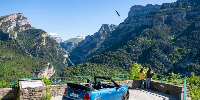 Deux femmes contemplent le canyon d'Anisclo depuis un mirador. Leur cabriolet est garé juste derrière et dans le ciel un vautour plane.