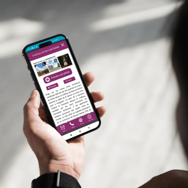 Une personne tient son smartphone avec l'application My Haute-Saône ouverte sur la page du Château de Ray-sur-Saône avec la possibilité de partager ses propres photos