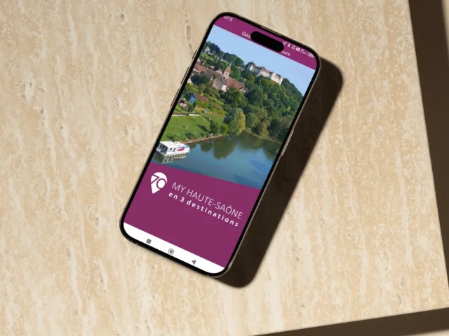 Un smartphone posé sur une table est ouvert sur l'application My Haute-Saône. On voit l’écran de bienvenue, où l’on aperçoit le château de Ray-sur-Saône, en Vesoul-Val de Saône, surplombant la Saône - Haute-Saône