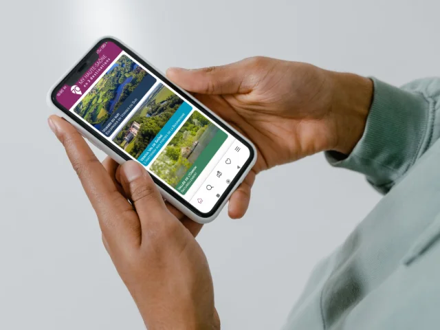 Une personne tient un smartphone avec l'application My Haute-Saône ouverte sur la page d’accueil, présentant les 3 destinations : Vesoul-Val de Saône, Vosges du Sud et Vallée de l’Ognon - Haute-Saône