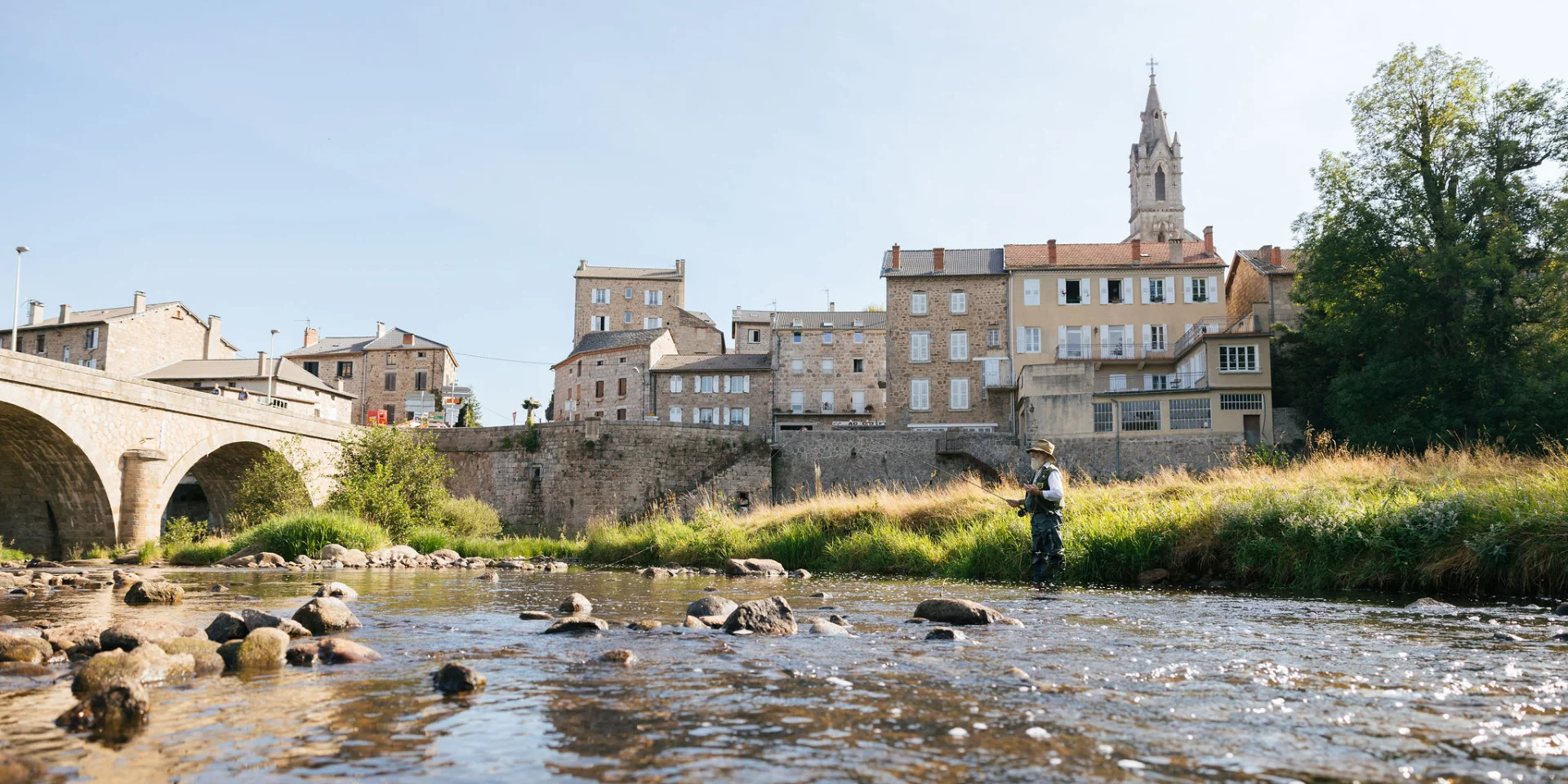 Au premier plan, on voit la rivière du Lignon qui coule avec un pêcheur à la mouche. Au second plan, un pont enjambe la rivière et les maisons alignées du village de Tence apparaissent serrées les unes contre les autres