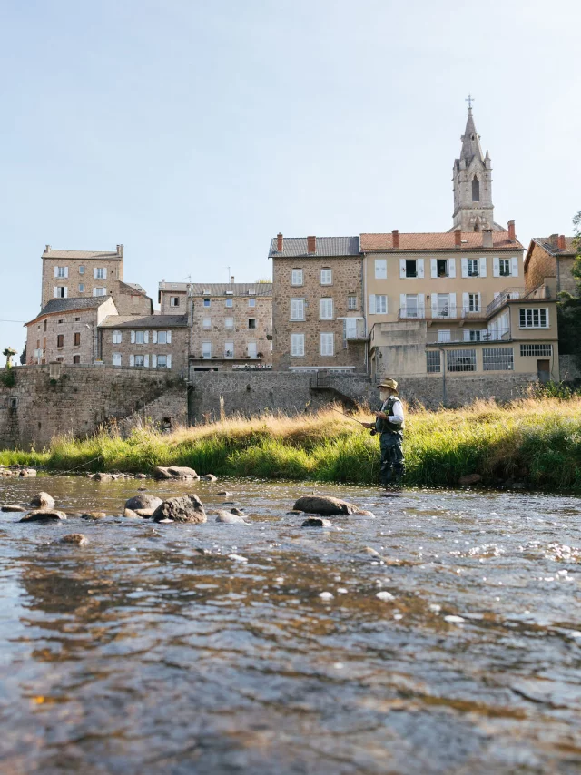 In the foreground, the river Lignon flows by with a fly fisherman. In the background, a bridge spans the river, and the houses of the village of Tence are tightly packed together.