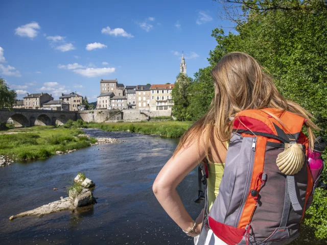 In the foreground, a Saint Jacques pilgrim looks out over the old village of Tence with its bridge and the Lignon river.