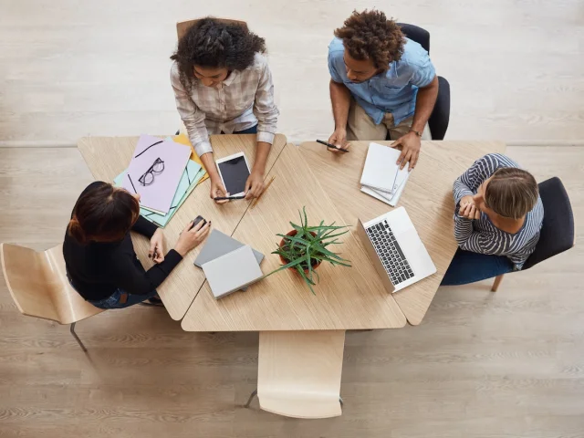 View from above of group young professional entrepreneurs sitting at table in coworking space, discussing profits of last team project, using laptop, digital tablet and smartphone