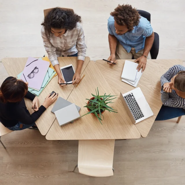 View from above of group young professional entrepreneurs sitting at table in coworking space, discussing profits of last team project, using laptop, digital tablet and smartphone