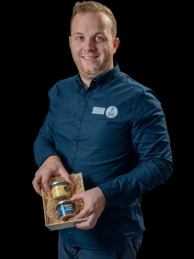 Sylvain Le Roux souriant, vêtu d'une chemise bleu et tenant dans ses mains deux pots de rillettes, sur fond noir