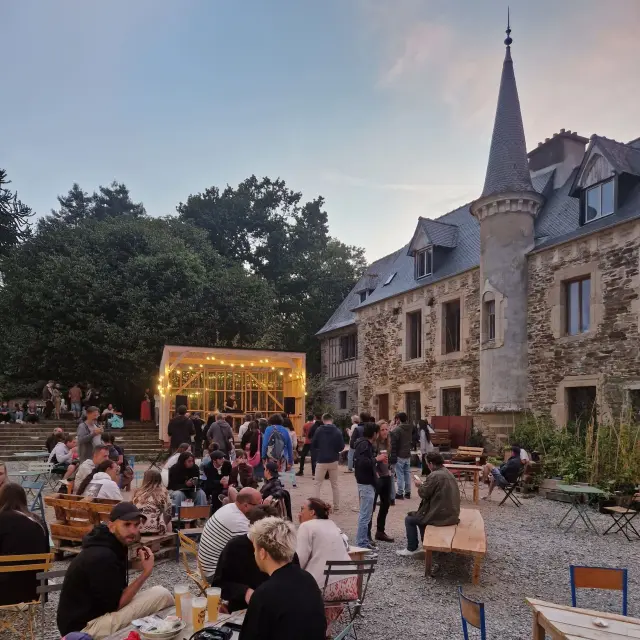 Des personnes assisses à table écoutent un concert dans la cours d'un manoir.